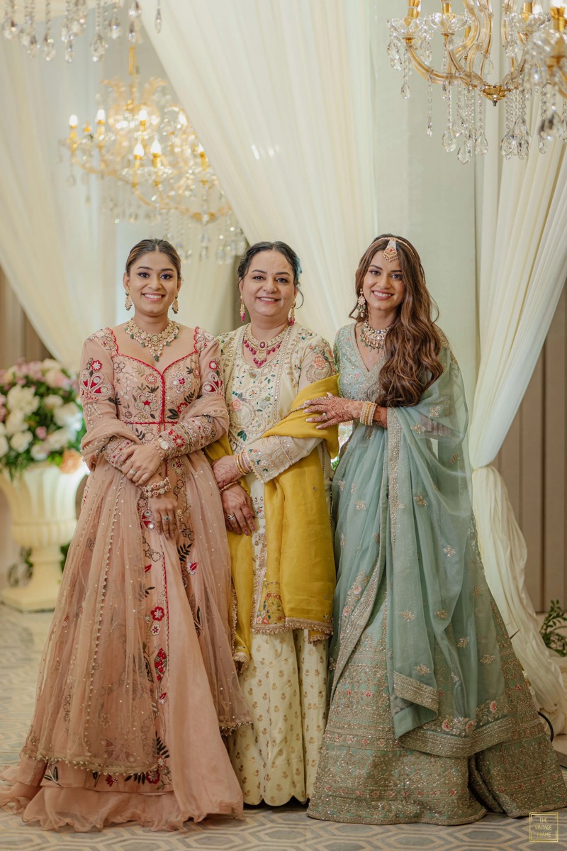 Three women dressed in elegant traditional Indian attire standing together at a festive event, with chandeliers and draped curtains in the background.