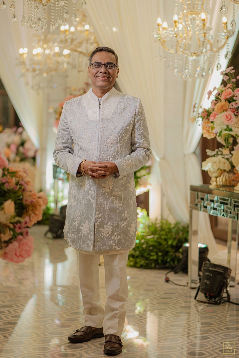 A man dressed in cream and gold traditional Indian attire standing in an elegantly decorated setting with chandeliers and floral arrangements.