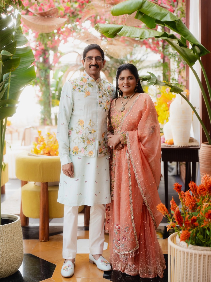 A man and woman in traditional South Asian attire standing together in a decorated setting with plants and flowers. The man is wearing a white embroidered kurta with matching shoes and white pants, and the woman is dressed in a peach saree with intri