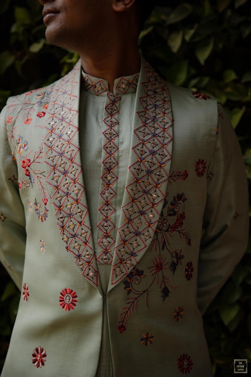 Person wearing a traditional embroidered jacket with floral and geometric patterns in various colors, standing outdoors against a leafy background.