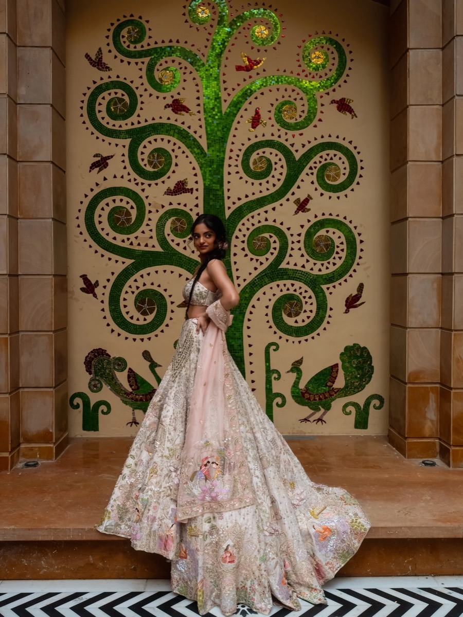 A woman in a traditional, detailed dress standing in front of a colorful mosaic mural of a green, curly tree with birds and peacocks.