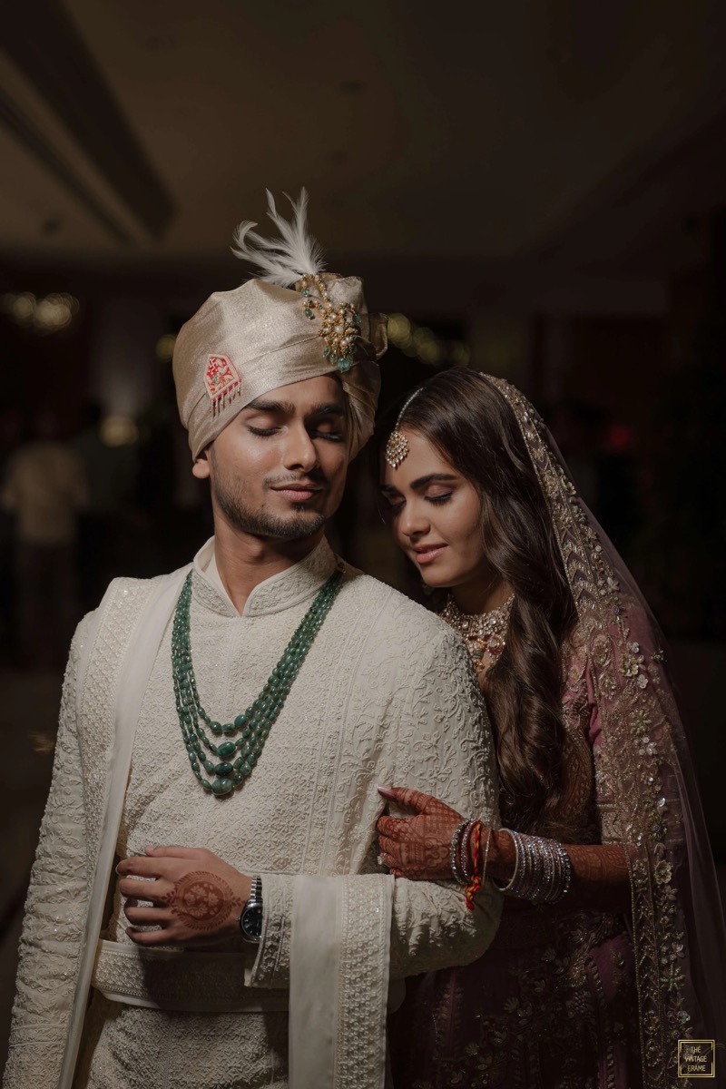 A newlywed Indian couple dressed in traditional attire, with the bride placing her hand on the groom's arm, at their wedding celebration.