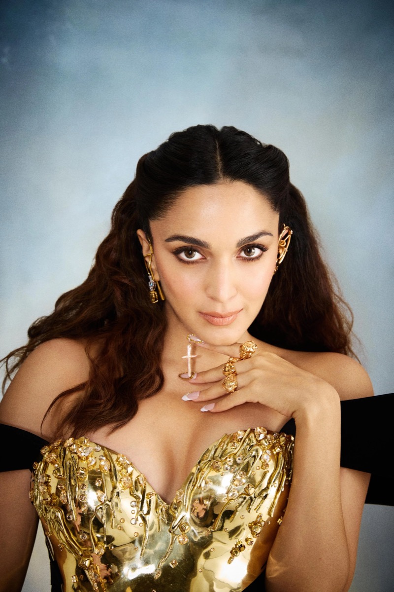 A woman with long, wavy brown hair, wearing gold jewelry including earrings, rings, and a necklace, poses against a blue-gray background. She wears a glamorous gold dress covered with embellishments and looks directly at the camera.