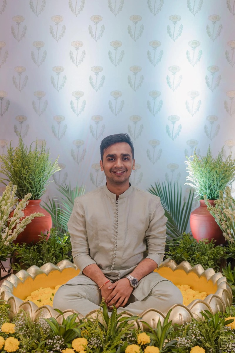 A young man sits cross-legged in front of a decorative backdrop with plants and flowers. He is smiling, wearing a light beige traditional outfit with a watch on his left wrist and a red string bracelet on his right wrist. The background features a pa