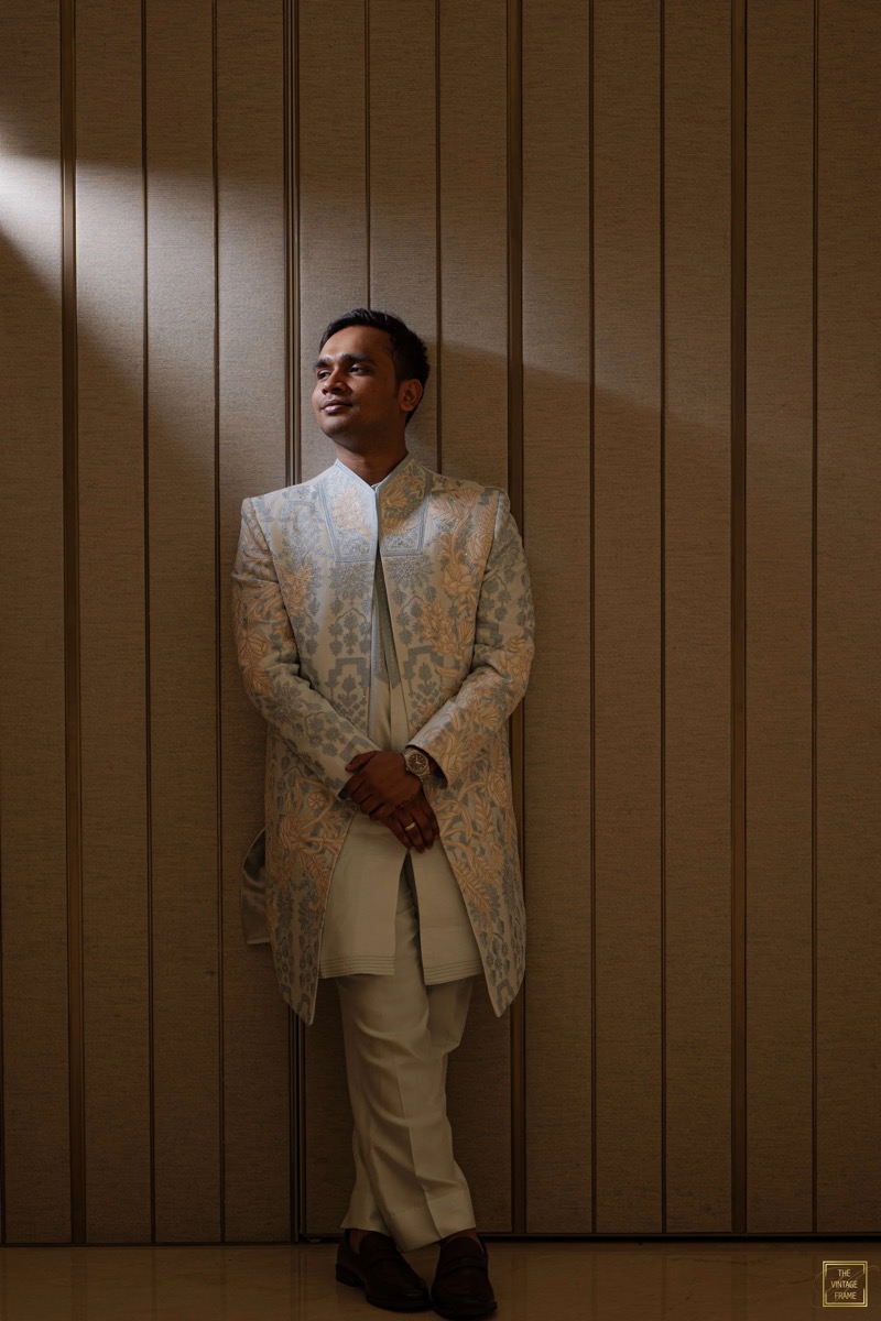 A man in traditional Indian attire, standing against a wooden-paneled wall, looking to the side with his hands clasped in front of him.