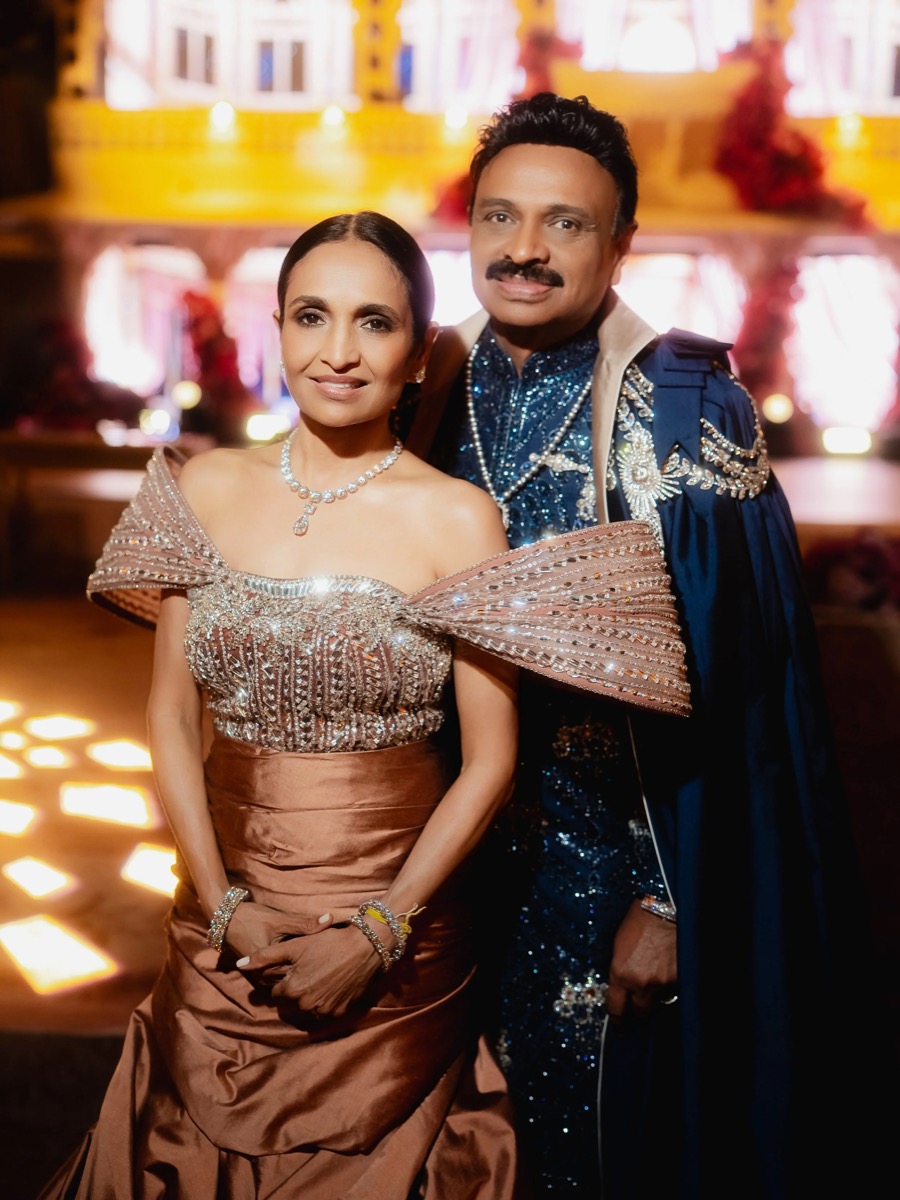 A couple dressed in elaborate, formal attire posing indoors with a decorative background.