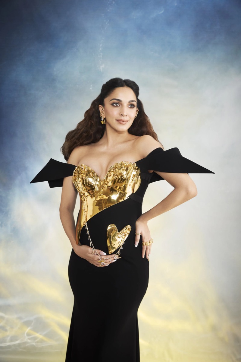 A woman wearing a black dress with a gold embellishment featuring a swan and a heart, standing against a cloudy sky background.
