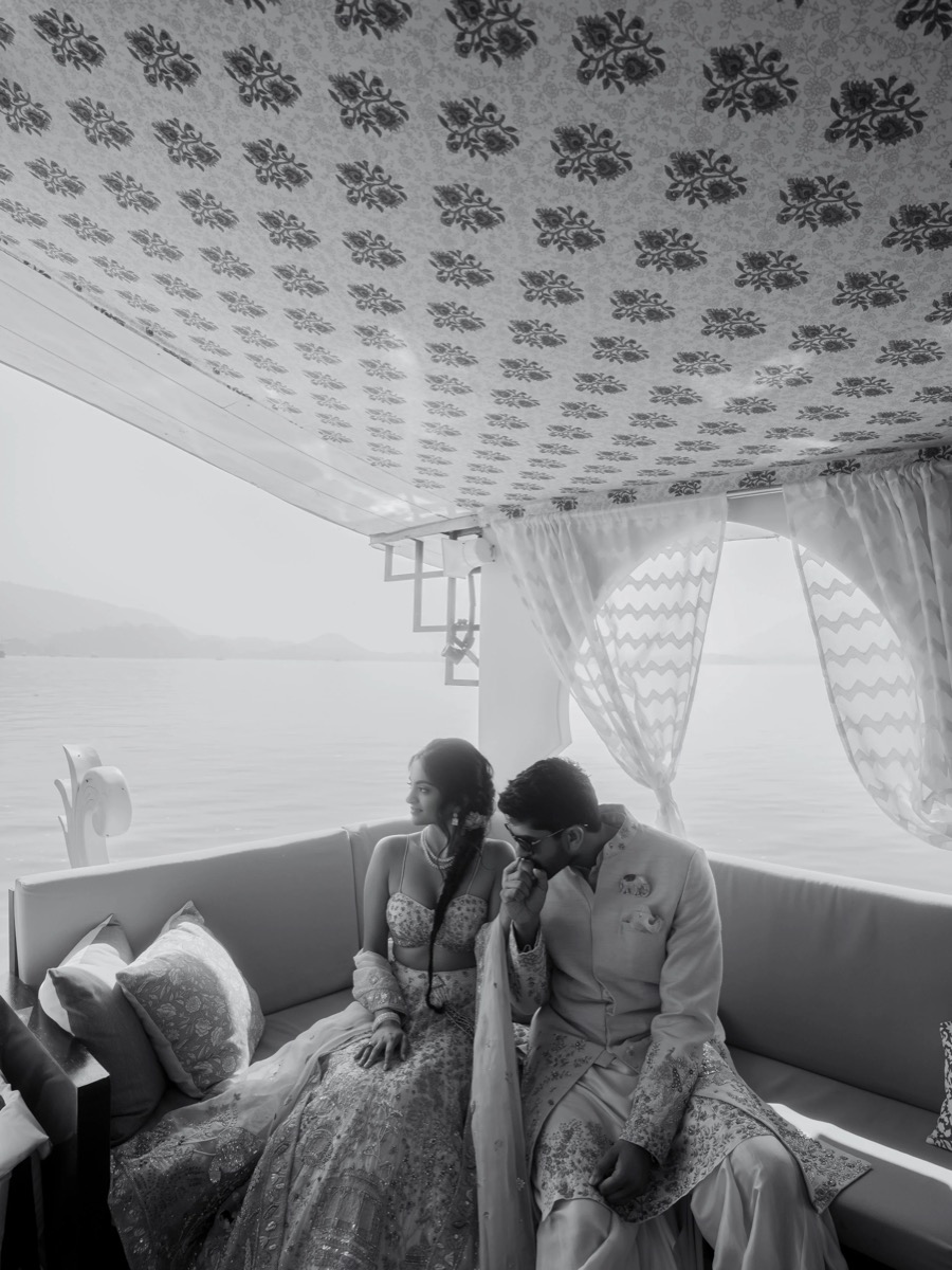 A black-and-white photo of a couple sitting on a sofa near a large window with curtains, by the water. The woman is dressed in traditional Indian attire and jewelry, while the man wears a sherwani, both appearing to be dressed for a wedding or specia