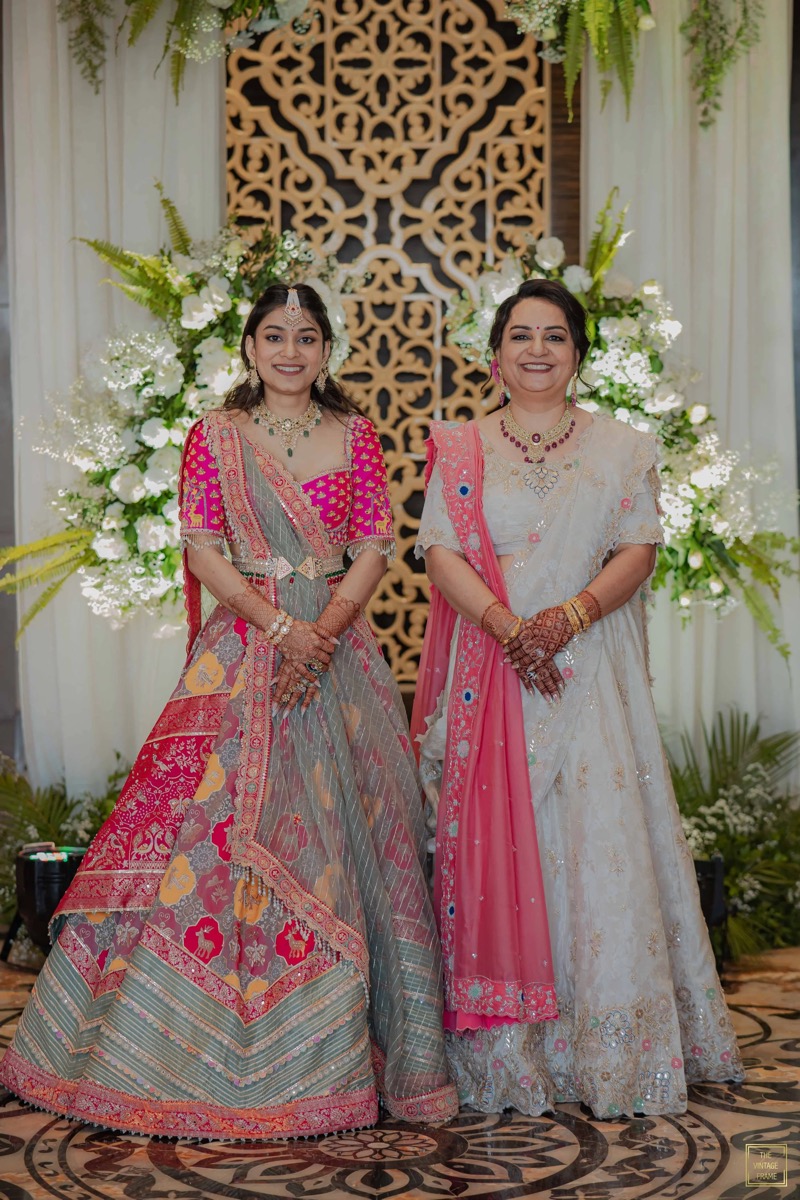 Two women dressed in traditional Indian sarees standing in front of decorated floral backdrop.