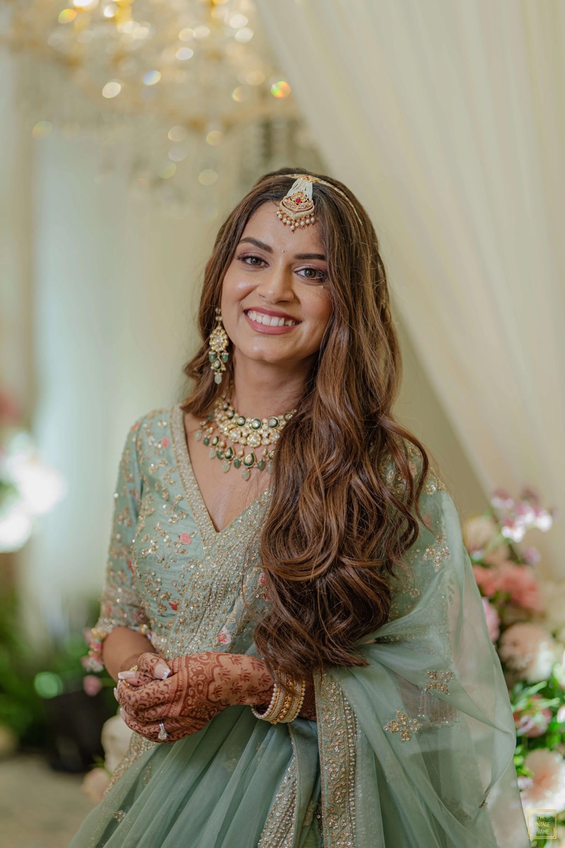A woman dressed in traditional Indian attire, wearing intricate jewelry, and standing in a decorated venue, smiling at the camera.