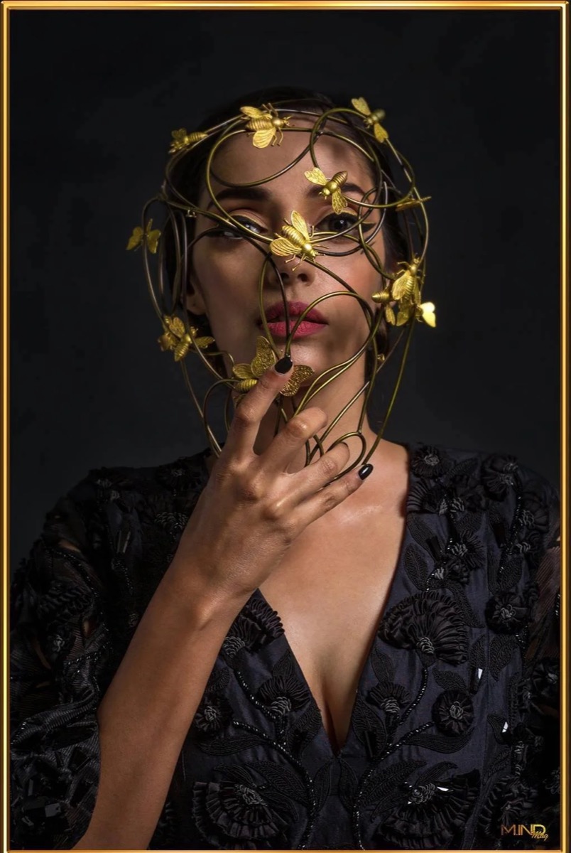 A woman is wearing a large, intricate headpiece made of gold-colored wire with numerous golden bee ornaments. She has a confident expression, with expertly applied makeup, and is dressed in a black embroidered top against a dark background.