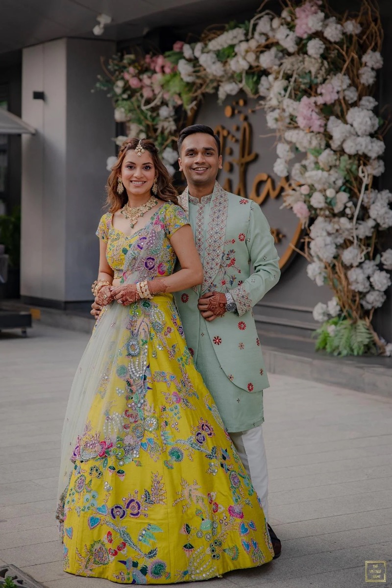 A couple dressed in traditional Indian attire stands in front of a floral backdrop, smiling at the camera. The woman wears a colorful, embroidered yellow gown with jewelry, and the man wears a light green embroidered kurta with white pants.
