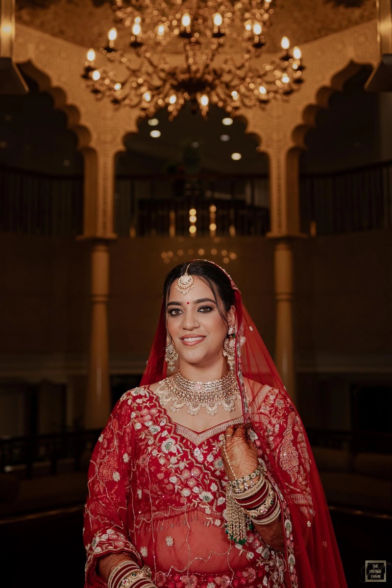 A smiling woman dressed in a traditional red bridal outfit with intricate embroidery and silver jewelry, standing in an ornate hall with a grand chandelier overhead.