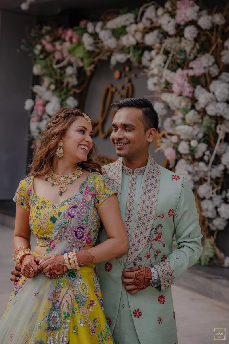 A smiling Indian couple in traditional attire at a festive event, surrounded by floral decorations.