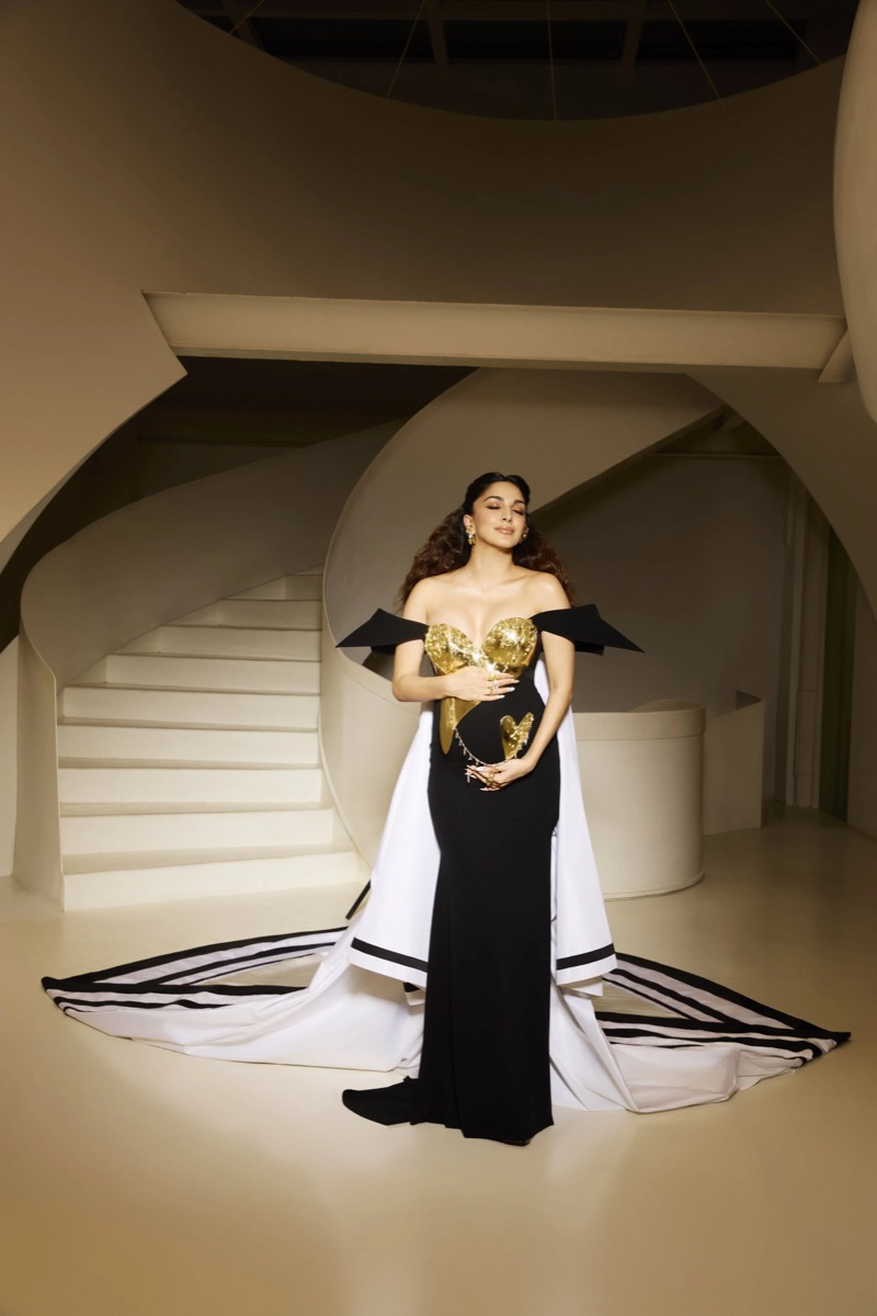Woman in a black and white gown holding golden heart-shaped decorations in front of a modern staircase and curved architectural background.