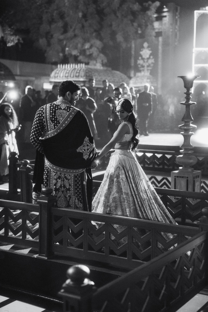 Black and white photo of a bride and groom holding hands during a wedding ceremony, surrounded by guests, with decorative umbrellas and lanterns in the background.