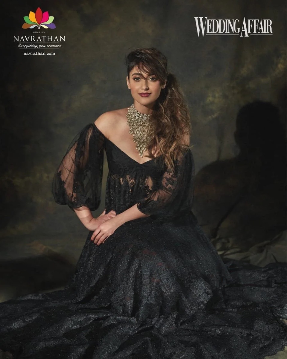 A woman with long, wavy hair and dark lipstick, wearing a black lace off-shoulder gown with sheer sleeves and a large Statement necklace, sitting against a dark backdrop.