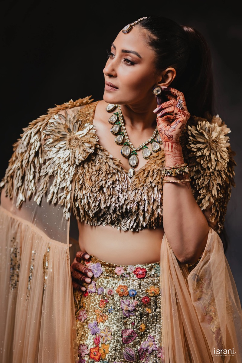 Woman wearing a beige embellished outfit with floral details, gold jewelry, and henna tattoos on her hand, posing against a dark background.