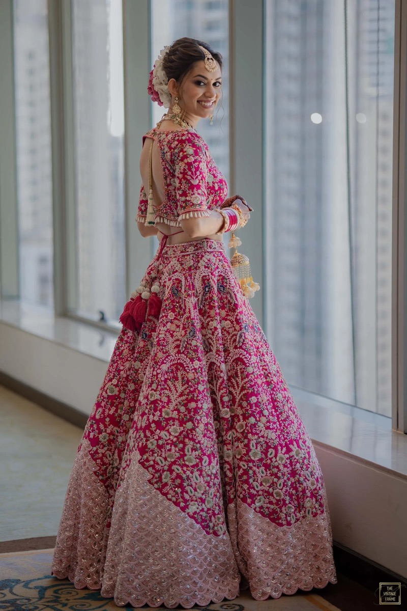 A woman dressed in a traditional pink and gold embroidered lehenga, smiling while standing by a window.