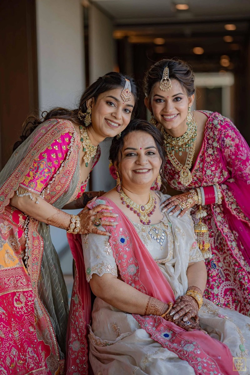 Three women in traditional Indian attire, jewelry, and henna, smiling and posing indoors, likely at a festive occasion.