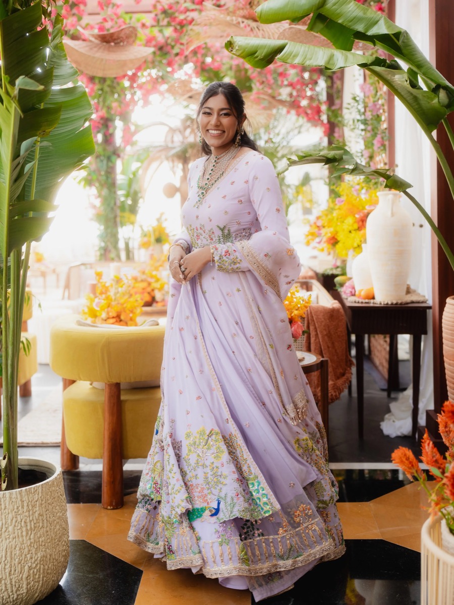 A woman dressed in a lavender embroidered traditional outfit standing in a decorated indoor space with lush green plants, colorful flowers, vases, and furniture.
