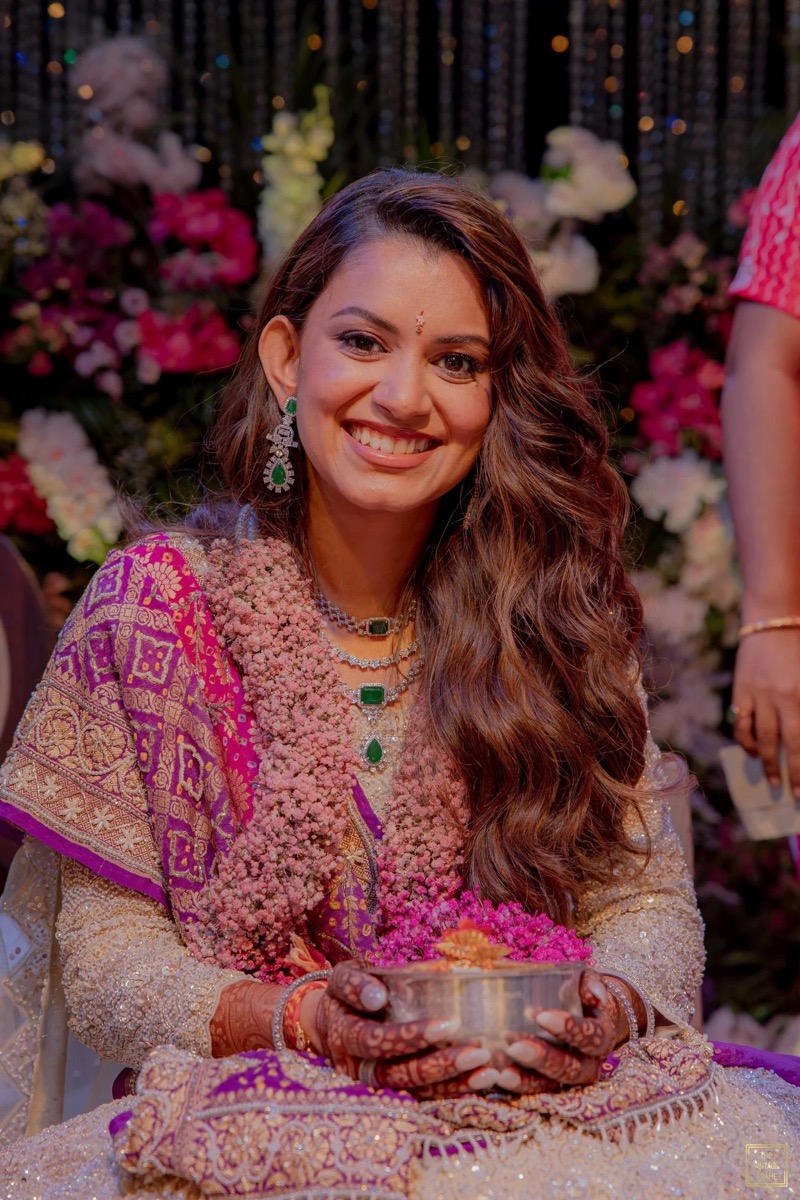 A smiling woman dressed in traditional Indian attire with jewelry and henna on her hands, holding a bowl decorated with flowers, during a festive celebration with floral decorations in the background.