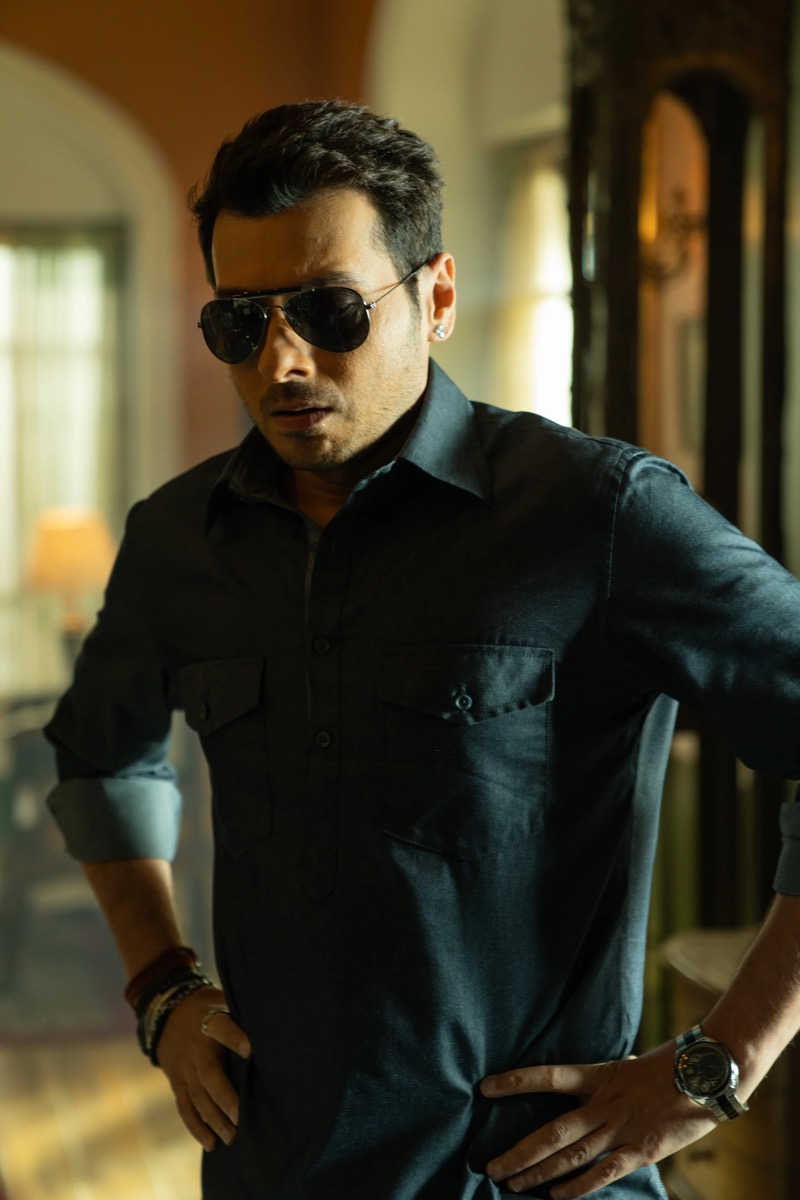 Man wearing black shirt, sunglasses, and watch standing indoors with hands on hips.