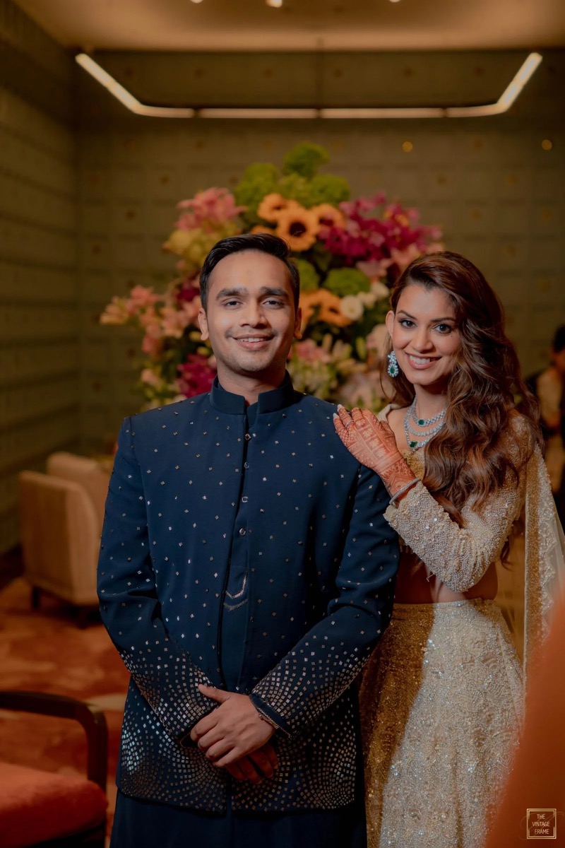 A man and woman dressed in traditional Indian wedding attire standing in a room with a large floral arrangement in the background. The man is wearing a dark blue kurta with gold accents, and the woman is wearing a gold-colored bridal outfit with jewe