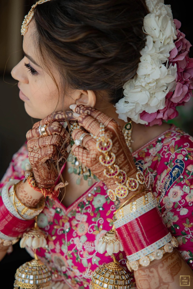 A woman dressed in traditional Indian attire, wearing elaborate gold jewelry and henna designs on her hands, adjusting her earring. Her hair is decorated with white and pink flowers.