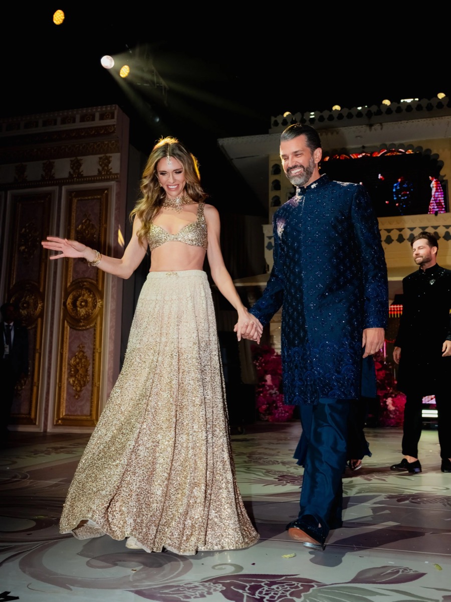 A man and woman dressed in traditional South Asian attire dance hand in hand on a decorated stage with ornate panels and floral arrangements.