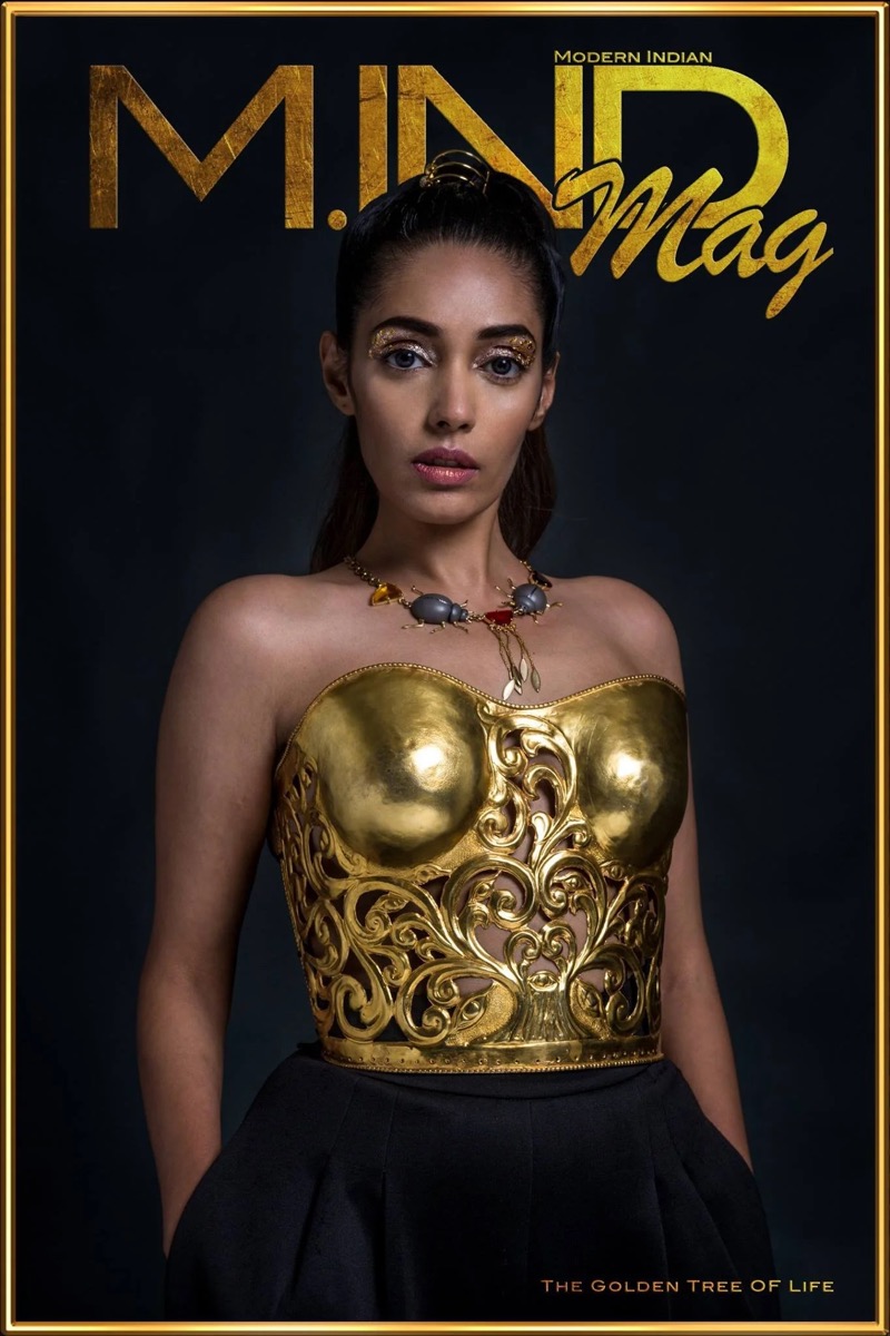 Magazine cover titled 'MIND Mag' featuring a woman with dark hair styled in a high ponytail, wearing gold eye makeup, a gold metallic strapless top with intricate swirling patterns, a colorful beaded necklace, and dark pants, against a dark backgroun