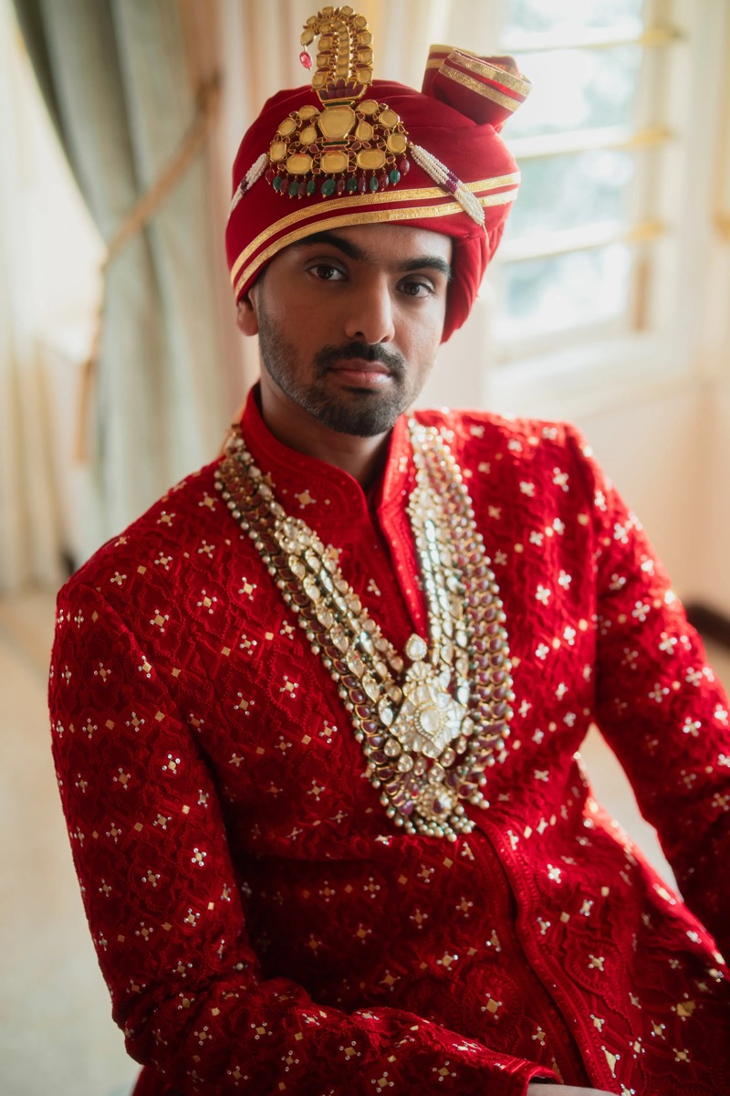 A man dressed in traditional Indian wedding attire, wearing a red embroidered sherwani, a red turban with gold embellishments and a peacock design, and multiple layered pearl and gemstone necklaces.