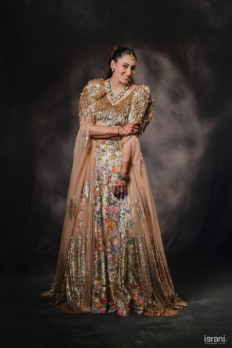 A woman wearing an ornate beige and gold embroidered gown with a sheer overlay, adorned with jewelry including earrings, necklaces, and a headpiece, standing against a dark background.