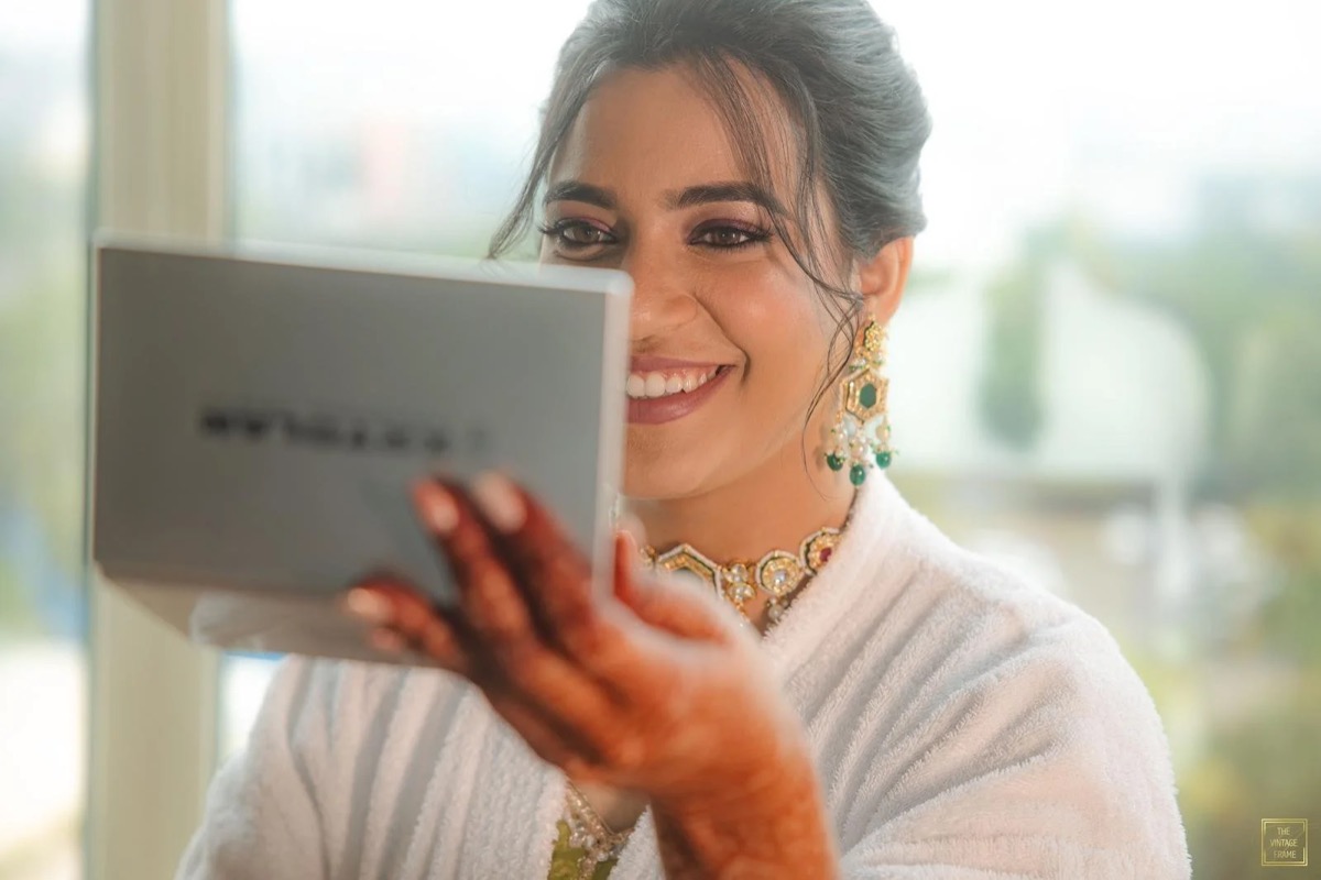 A woman with traditional Indian jewelry and henna on her hand, smiling and looking into a mirror.