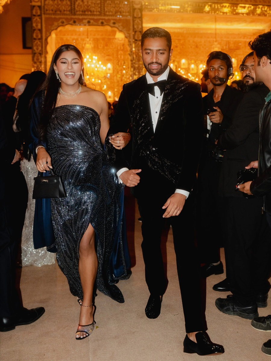 A man and woman in formal attire walking hand in hand at an event with a decorated background and other guests in the background.