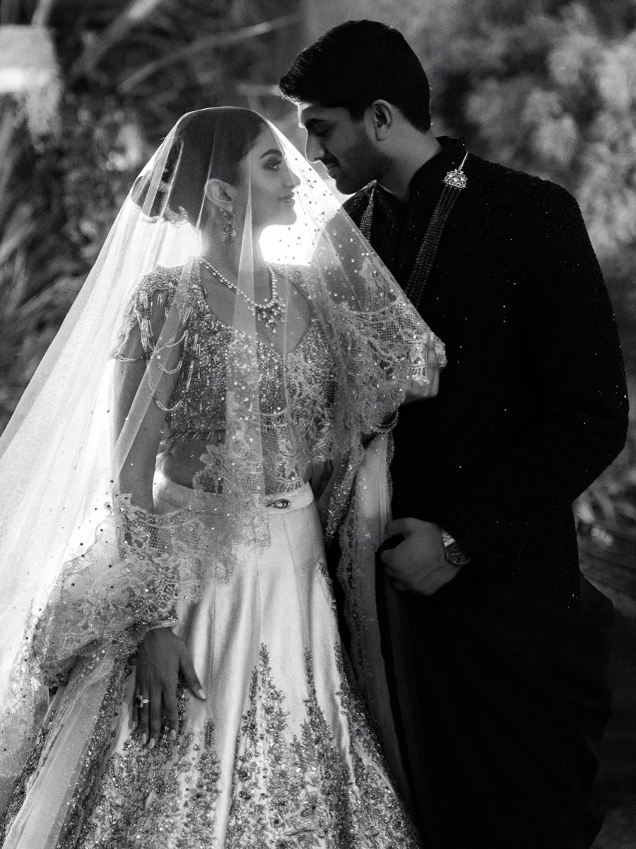 A black and white photo of a bride and groom standing close together, gazing at each other. The bride is wearing an ornate traditional dress with a veil, and the groom is dressed in a dark, decorative suit. They are outdoors with blurred natural back