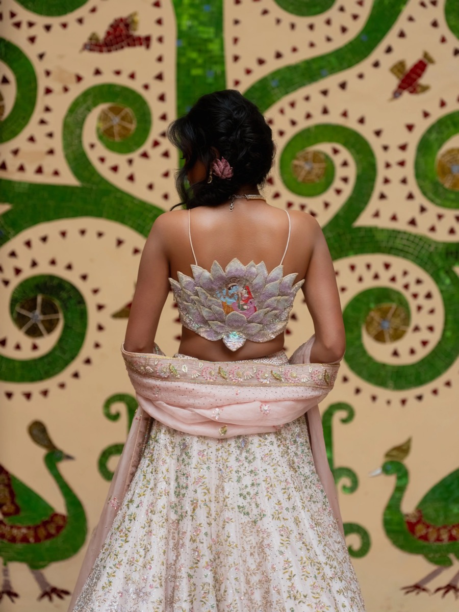 Back view of a woman in traditional Indian attire, standing in front of a decorative background with green and cream swirls and peacock motifs.