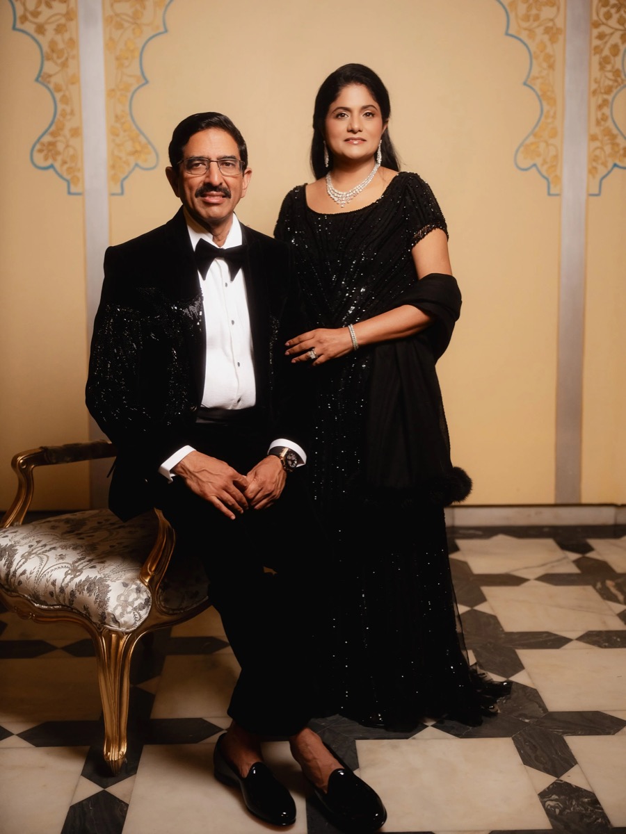 A man in a tuxedo sitting on a fancy chair and a woman in a black evening gown standing next to him, both looking at the camera, in a decorated indoor setting.