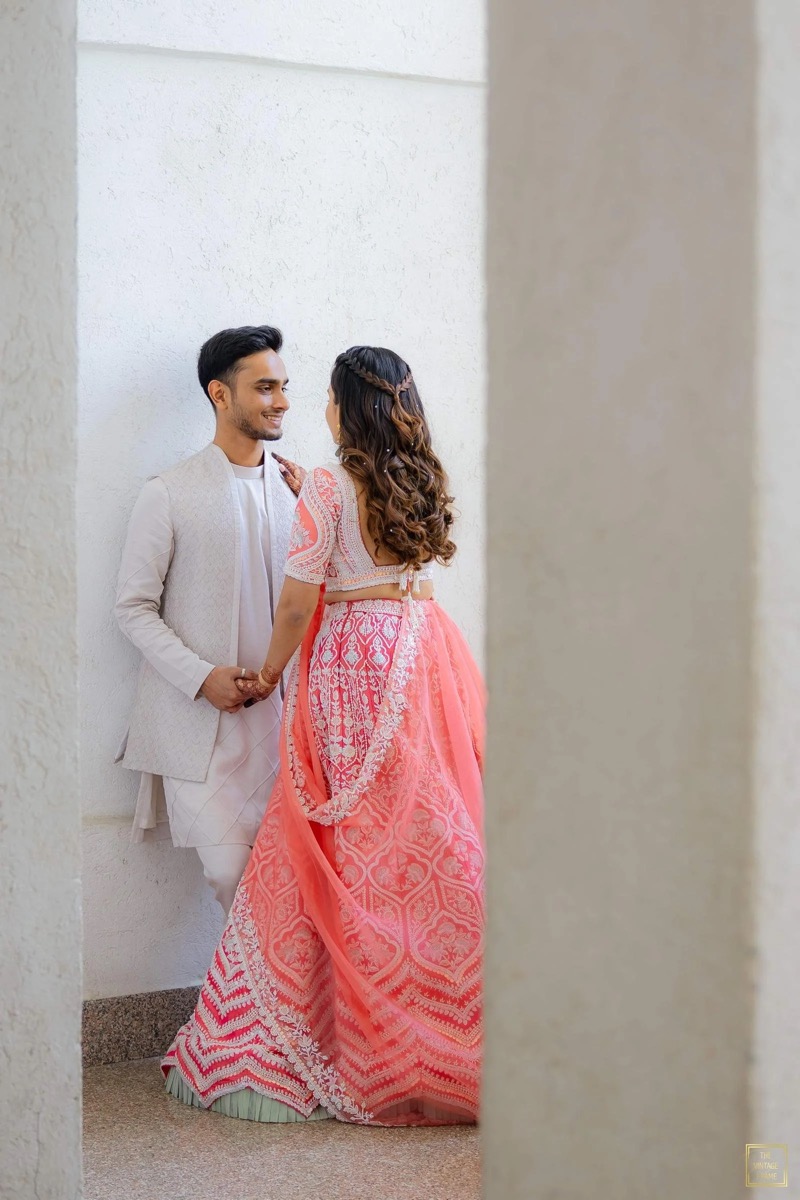 A young couple dressed in traditional Indian clothing, standing close and smiling at each other against a plain white wall, with beige-colored pillars partially framing the scene.