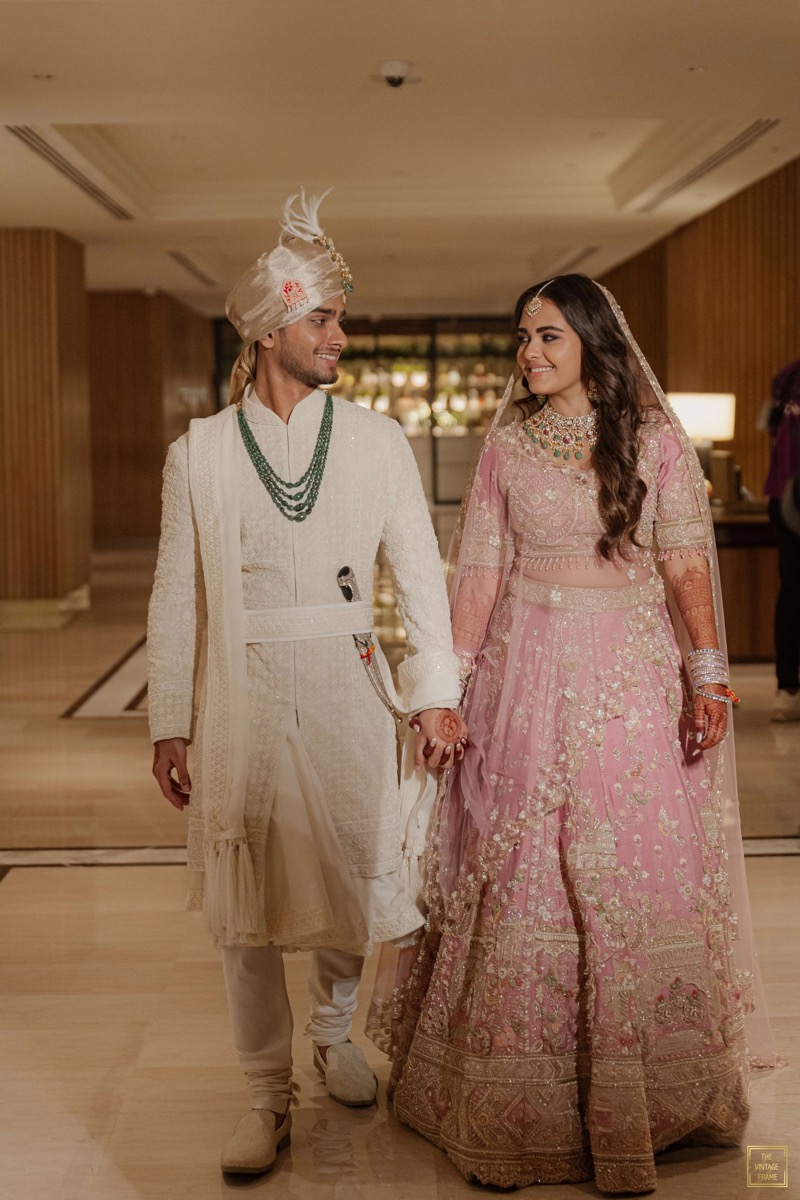 A bride and groom holding hands, dressed in traditional Indian wedding attire, walking indoors with wooden walls and warm lighting.