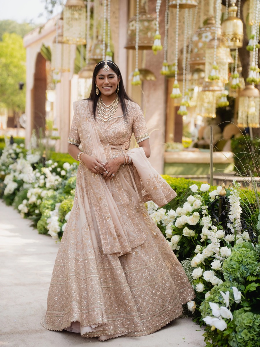 A woman dressed in an elegant beige traditional Indian outfit with intricate embroidery, standing outdoors near a decorated aisle with white flowers and hanging bells, smiling at the camera.