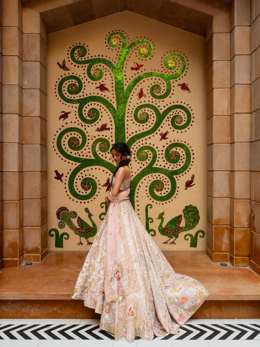A woman in a fancy embroidered gown standing in front of a large colorful mosaic wall featuring a green tree with swirling branches, flowers, and birds.