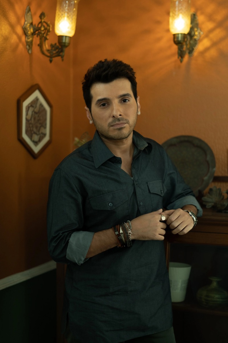 A man with dark hair and light facial hair, wearing earrings, a dark button-up shirt with rolled-up sleeves, multiple bracelets, and a watch, stands indoors against a background of orange walls with framed artwork and ornate wall sconces, looking dir