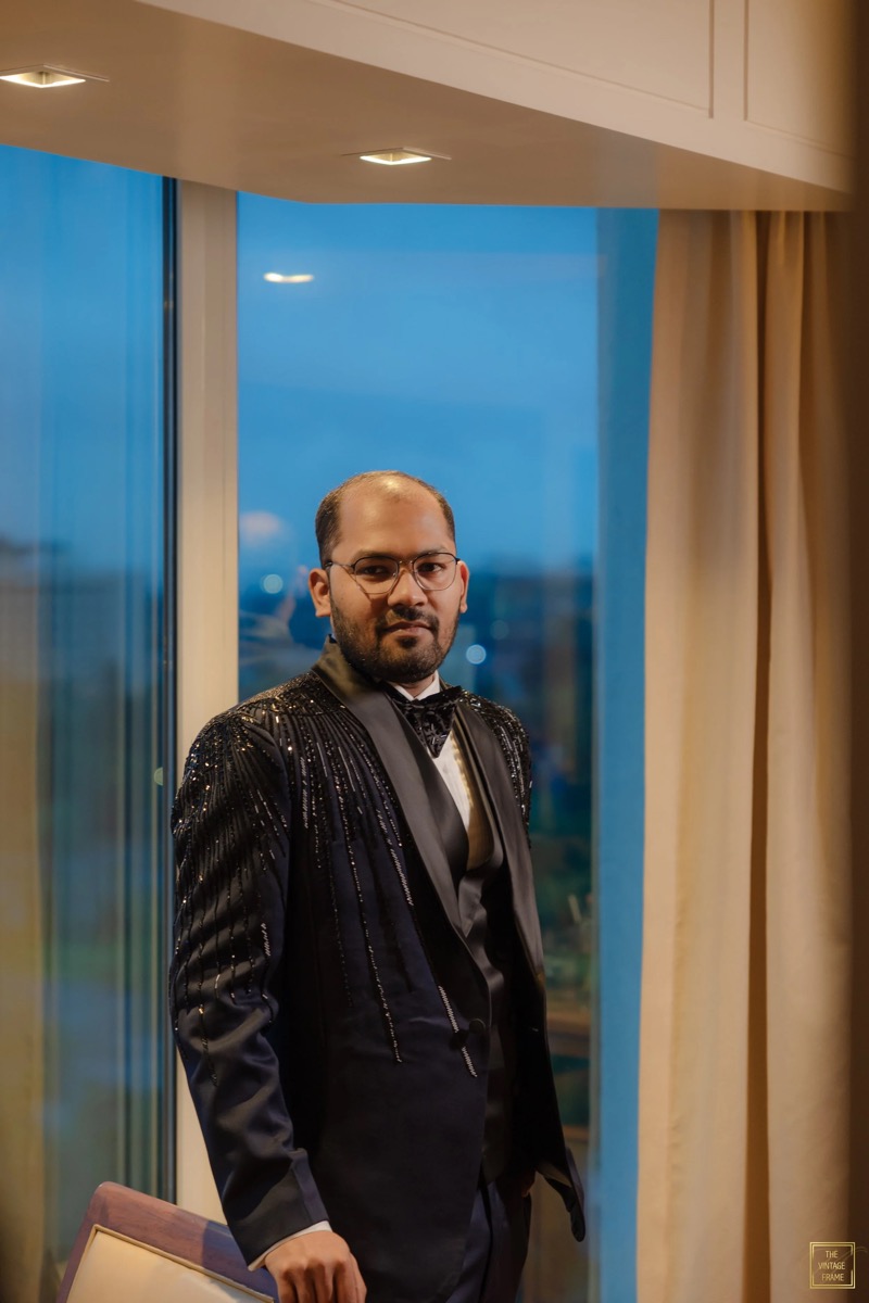 A man dressed in a black tuxedo with embellishments, standing indoors near a window with beige curtains, during evening or night.