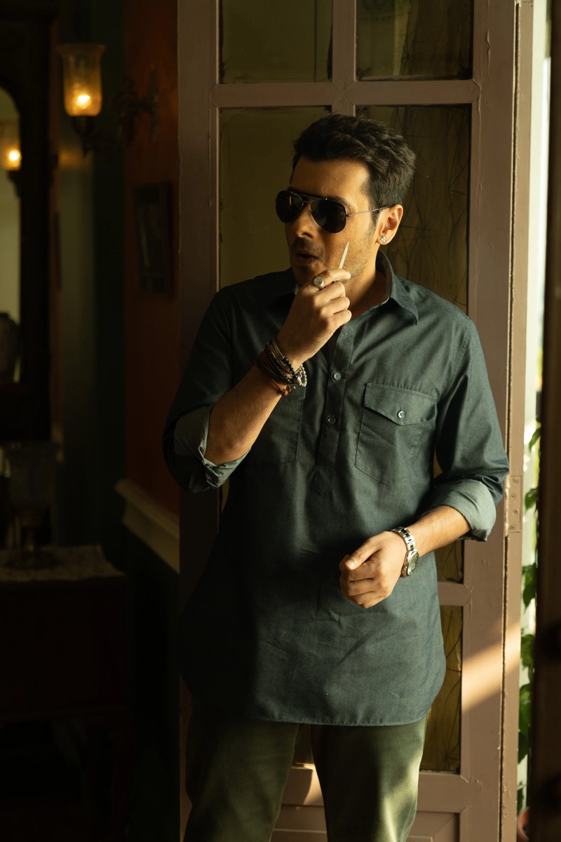 A man standing indoors, wearing sunglasses, a dark button-up shirt, and various jewelry, holding a toothpick near his mouth, and looking to the side near a window.