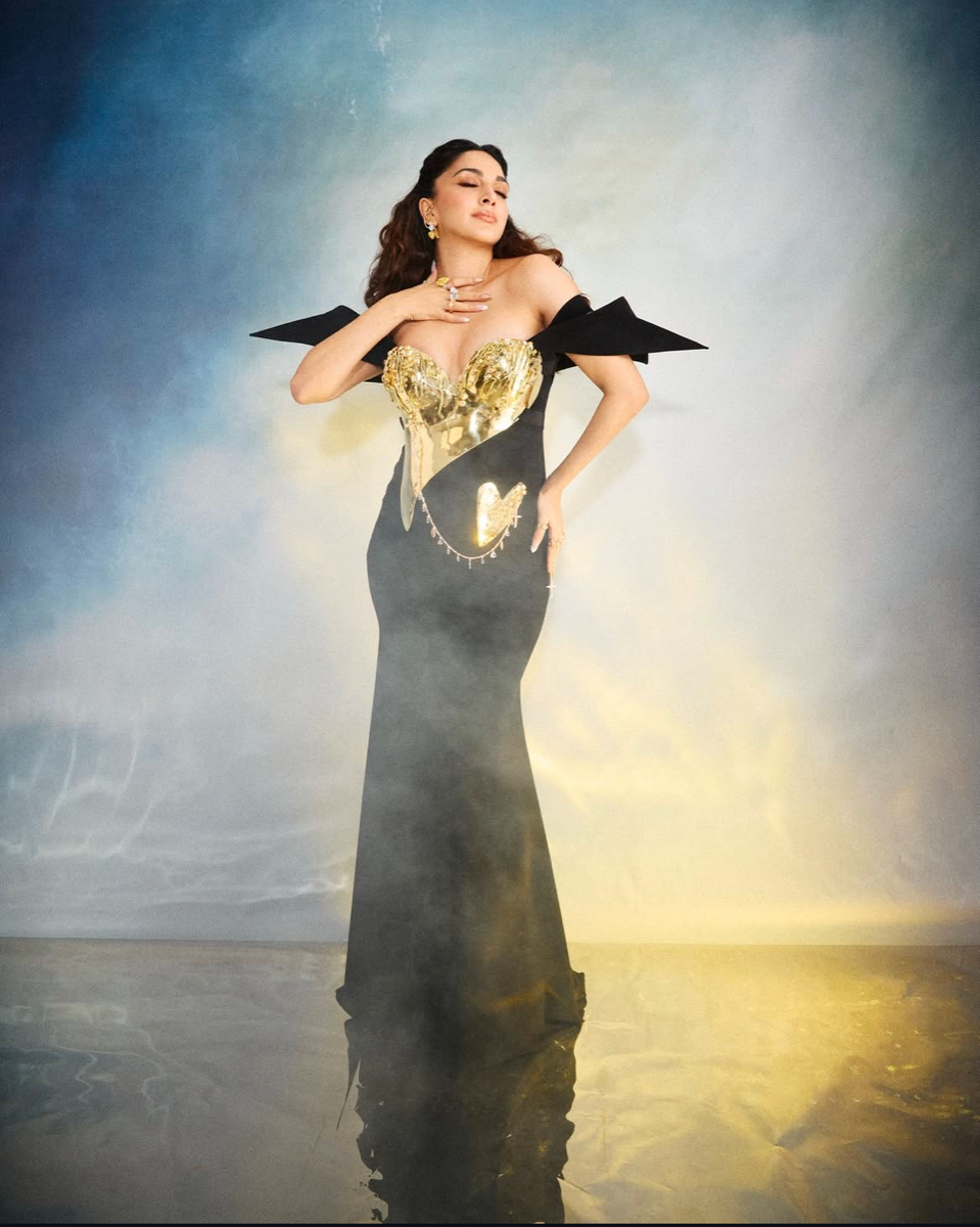 A woman in a long black dress with a gold heart-shaped bodice and shoulder accents, standing in shallow water with a cloudy sky background, posing with her eyes closed and one hand on her chest, the other on her hip.