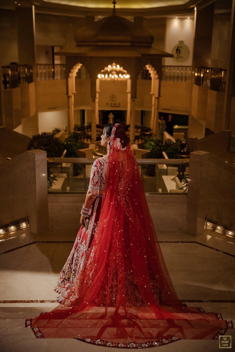 A bride dressed in a red traditional Indian wedding outfit with a long veil, standing on a staircase inside a grand, dimly lit building with ornate decor and chandeliers.