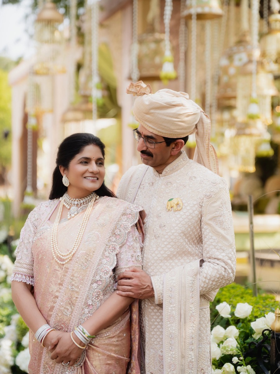 A couple dressed in traditional Indian wedding attire standing close together, smiling at each other, surrounded by white flowers and decorative elements in the background.