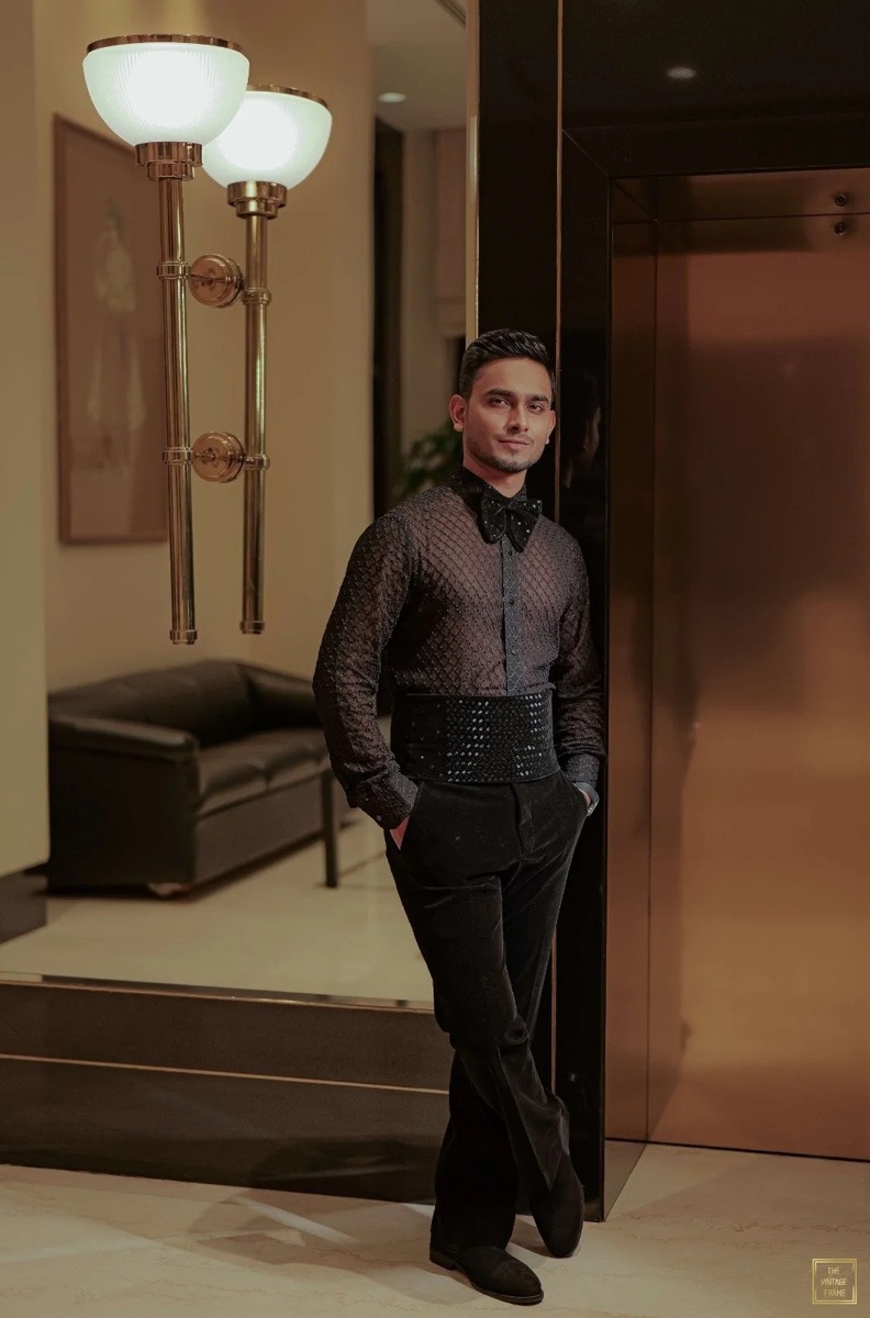 A man dressed in black formal wear, standing in front of an elevator, with hands in pockets in a hotel lobby.