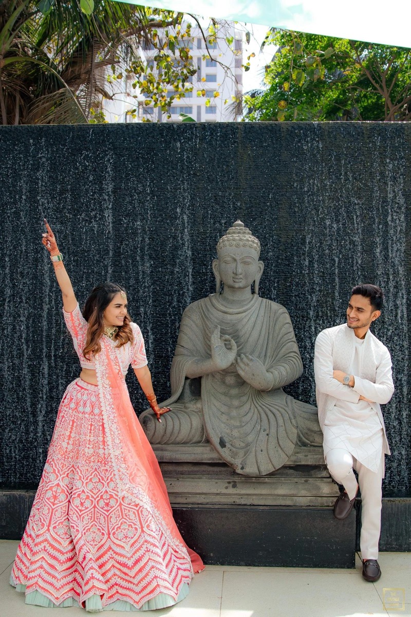 A woman in a pink traditional Indian dress and a man in white traditional attire are posing next to a seated Buddha statue with a waterfall in the background.